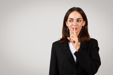 Playful businesswoman with a mischievous grin placing her finger over lips in a 'shush' gesture