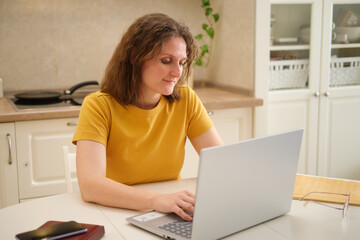 A woman with a laptop is typing on a keyboard while sitting at a table in a home kitchen. An adult female businesswoman works from home, a remote office