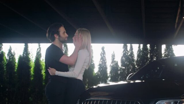 A couple in love is kissing while standing in front of their car. They are affectionate, hugging and touching each other. The man takes a woman&rsquo;s face in his hands and kisses her again.
