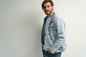 Fototapeta premium Portrait of stylish brutal young bearded European man wearing jeans jacket, posing at white blank wall with copy space for your text or promotional content