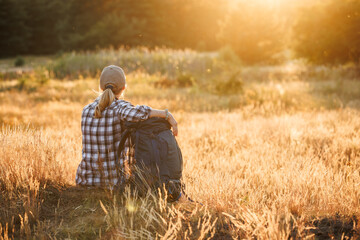 Woman with backpack is resting in nature and enjoying summer sunset. Relax while hiking on a long...
