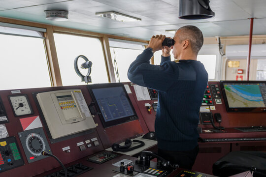 Officer on watch with binoculars on the navigational bridge. Caucasian man in blue uniform sweater on the bridge of cargo ship.