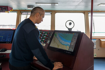 Officer on watch with electronic chart on the navigational bridge. Caucasian man in blue uniform sweater using ECDIS on the bridge of cargo ship.