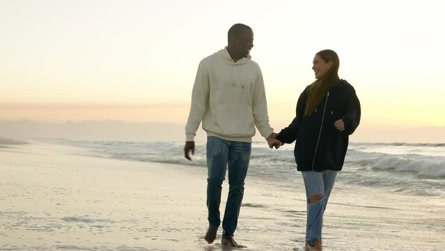 Casually Dressed Loving Couple Walking Through Waves Along Beach Shoreline Holding Hands At Sunrise