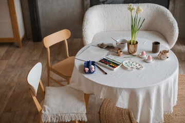 Close-up of a paintbrush, paint palette and carton of eggs being painted for Easter decorations with narcissus flowers on a table