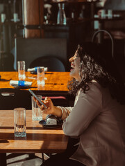 Young woman, laughing while she is holding her phone, She is in a fancy restaurant, waiting for service.