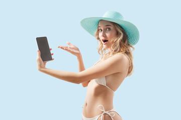 amazed woman dressed in a white bikini an big blue sun hat is showing a smartphone. Image is...