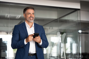 Smiling middle age entrepreneur using cell phone mobile app, looking aside. Digital technology application and solutions for business. Mature 40s businessman wearing suit holding smartphone indoors. 