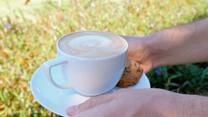 The person is holding a cup of coffee and a cookie on a saucer with their thumb and finger. Coffee on the grass background, relaxing moment of sharing food.