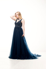 Fototapeta premium Young blonde woman in a dark blue ball gown with a fluffy skirt on a white background.