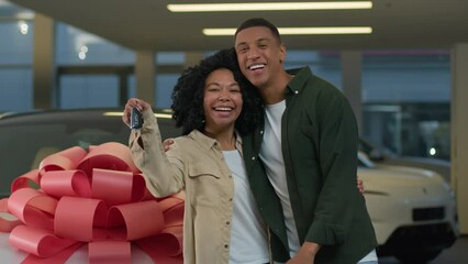 Excited young happy couple in love celebrating purchase new car luxurious present with big bow on automobile hood African American woman man showing keys of auto buying in modern dealership showroom