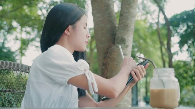 Young Asian woman at writing work noted down important ideas onto digital tablet, memorizing method, use portable working gadget, drawing on tablet screen, relaxing day at outdoor cafe coffee table