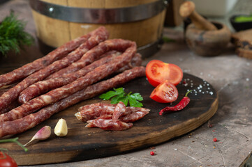 Rustic dry thin sausages among vegetables and herbs on a dark table