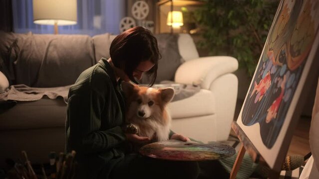 A beautiful young female artist is painting a picture in her cozy studio. A woman is petting her fluffy corgi dog while creating her work of art. Inspired girl illustrator working at home