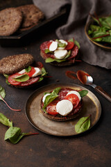 Rye bun sandwiches or burgers with salami, mozzarella, cherry tomatoes, chard leaves and ketchup on dark concrete background. Side view