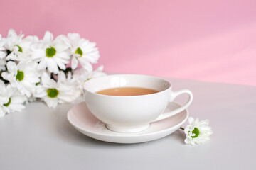 A white cup with tea and a saucer stands on a table with white chrysanthemum flowers