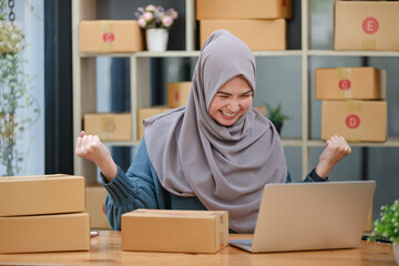 Active asian woman in blue muslim suit sitting and working with computer and online package box...