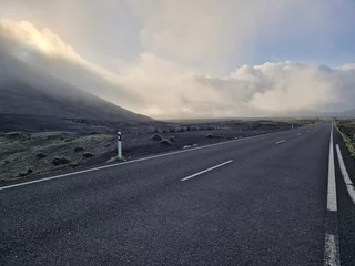 Papier Peint photo les îles Canaries Black asphalt road in black volcanic landscape of Lanzarote, with low hanging clouds in the background