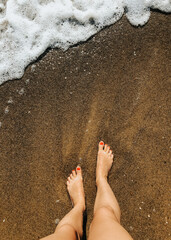 Woman legs barefoot at sea foam waves on sand beach summer day. top view above women feet.