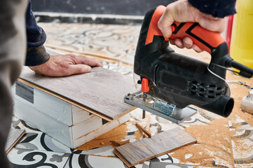 Close up of male construction worker cutting laminate wooden panel with jigsaw tool. Man using electric power saw cutter while preparing laminate for floor installation.