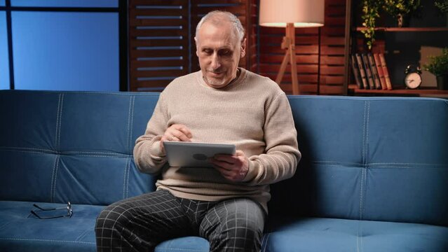 Elderly man using new tablet, grandfather sitting on couch alone at home in evening, portrait shot