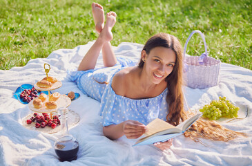 Picnic scene: pretty smiling girl lying on plaid and reading book in park. Outdoor portrait of...