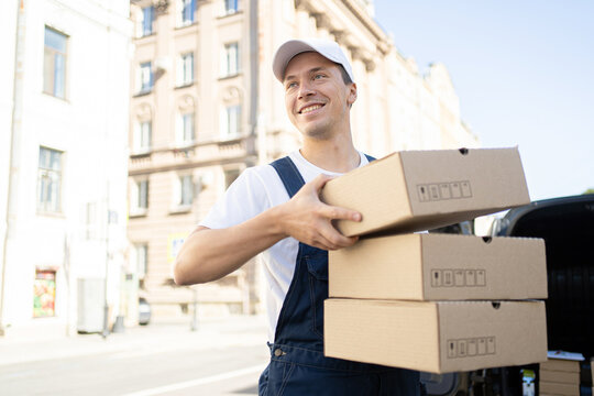A delivery man in a white cap and overalls happily carries cardboard boxes for delivery in an urban setting.