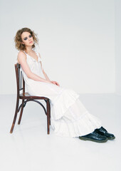 Fototapeta premium Portrait of pretty strange sad girl. Beautiful weird young woman in white dress sitting on stool in empty room