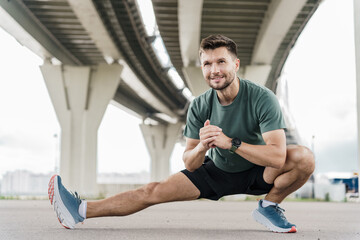 Focused man doing stretching exercises under an overpass, wearing a smartwatch and sporty attire.