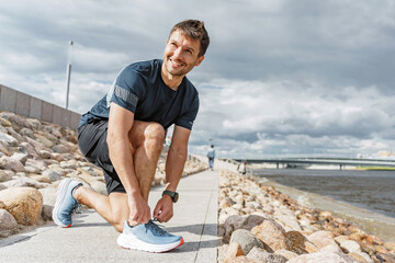Smiling runner tying shoelaces on a waterfront walkway, ready for a workout with a cityscape background.
