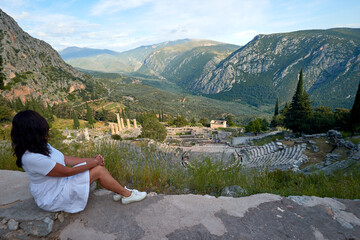 Greek history:  A long-haired woman from behind, wearing white dress  looking at Apollo Temple and Ancient Theater in sunset. Delphi, Greece.