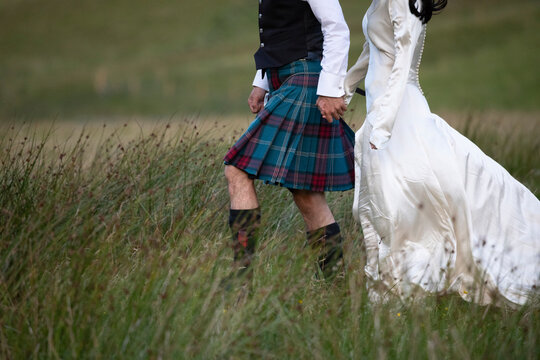 A newly married Scottish couple strolls through a grassy field in Glencoe, the Scottish highlands. He dressed in a Scottish Kilt and she dressed in a white wedding dress