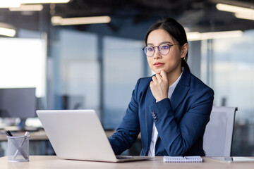 Professional young female executive deep in thought while working at a laptop in a contemporary office setting.