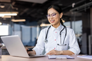 Smiling Asian female doctor in a white coat with stethoscope using laptop in a well-lit contemporary office setting.