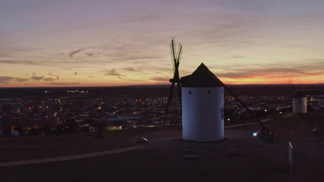 AERIAL VIEW OF A SPANISH OLD WINDMILL AT SUNSET WITH VILLAGE LIGHTS IN THE BACKGROUND