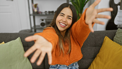 Young hispanic woman smiling in a cozy indoor home setting, reaching out as if capturing a selfie, portraying warmth and friendliness.