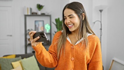 Smiling young hispanic woman takes a selfie in a stylish living room, wearing casual attire.