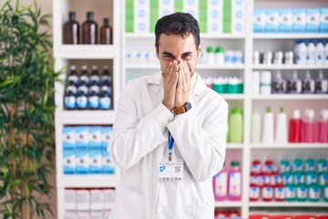 Handsome hispanic man working at pharmacy drugstore laughing and embarrassed giggle covering mouth...