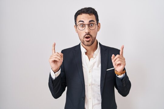 Handsome business hispanic man standing over white background amazed and surprised looking up and pointing with fingers and raised arms.