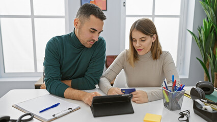 A woman and a man coworking in a bright office analyze data on a tablet, with stationery and a card visible.