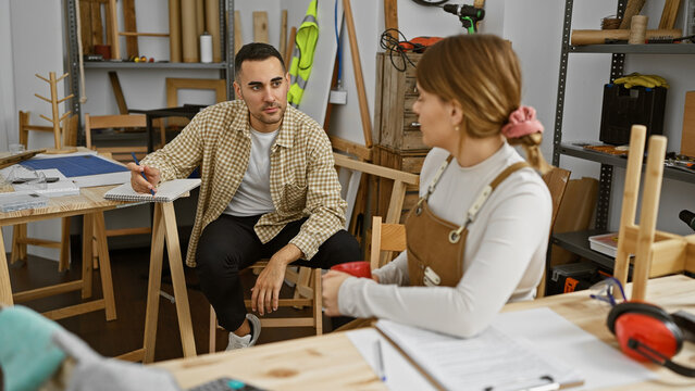A man and woman discuss work in a well-equipped carpentry workshop, surrounded by wooden furniture and tools.
