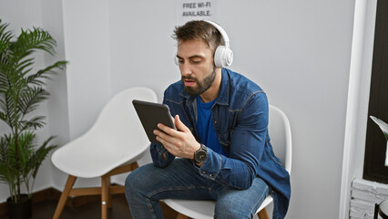 Relaxed young hispanic man enjoying tech-life in waiting room, sitting with touchpad, immersed in music through headphones