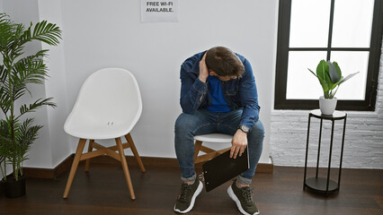 Stressed young hispanic man holding a clipboard, sitting restlessly in the waiting room, his...