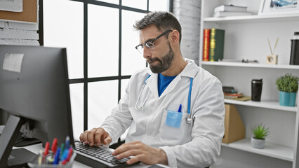 Fototapeta na wymiar Bearded young hispanic man doctor hard at work, using his computer indoors at a medical clinic