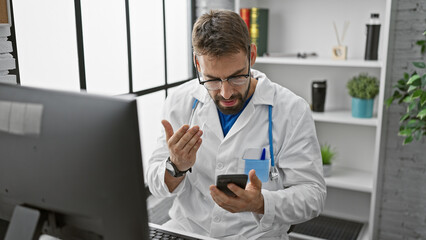 Serious young hispanic male doctor engrossed in typing a message on his smartphone, seated at the clinic's interior, textures of healthcare professionalism vividly painted in the background. - obrazy, fototapety, plakaty