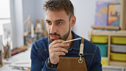 In the heart of the art studio, doubting yet determined, young hispanic man artist in an apron,...