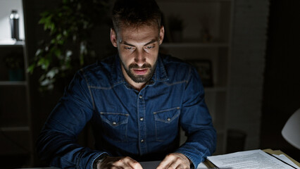 Obraz premium Driven young hispanic man, working long nights at office; focused, serious yet relaxed, this bearded professional navigates business on his laptop.