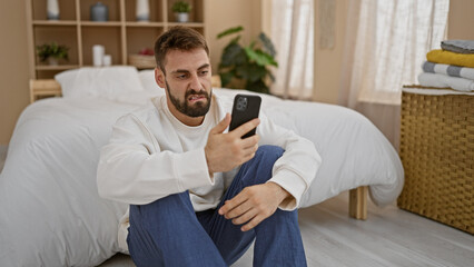 Handsome, young hispanic man chilling on the bedroom floor, lost in the online world on his phone,...