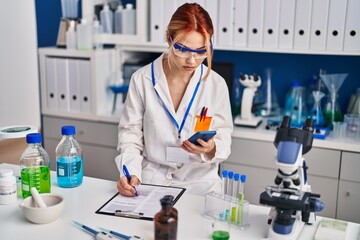 Fototapeta premium Young caucasian woman scientist using smartphone writing document at laboratory