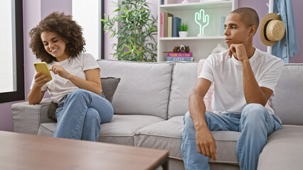 Beautiful couple unhappy at home, sitting on sofa. girlfriend happily texting, igniting jealousy. their love strained over smartphone use indoors.